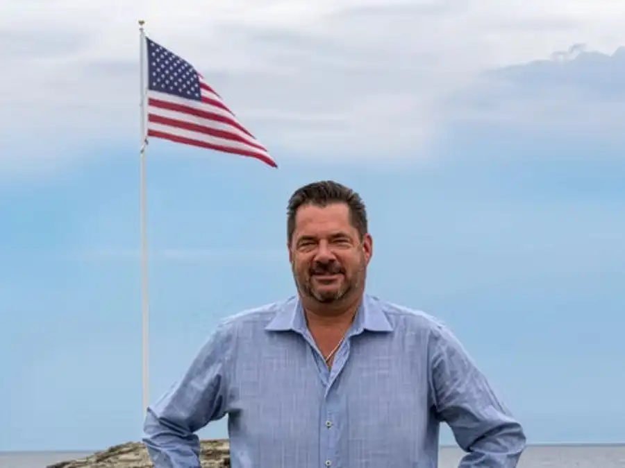 Candidate for State Representative, Ray Collins III, stands smiling outdoors with hands on hips, in front of an American flag and a waterfront view.