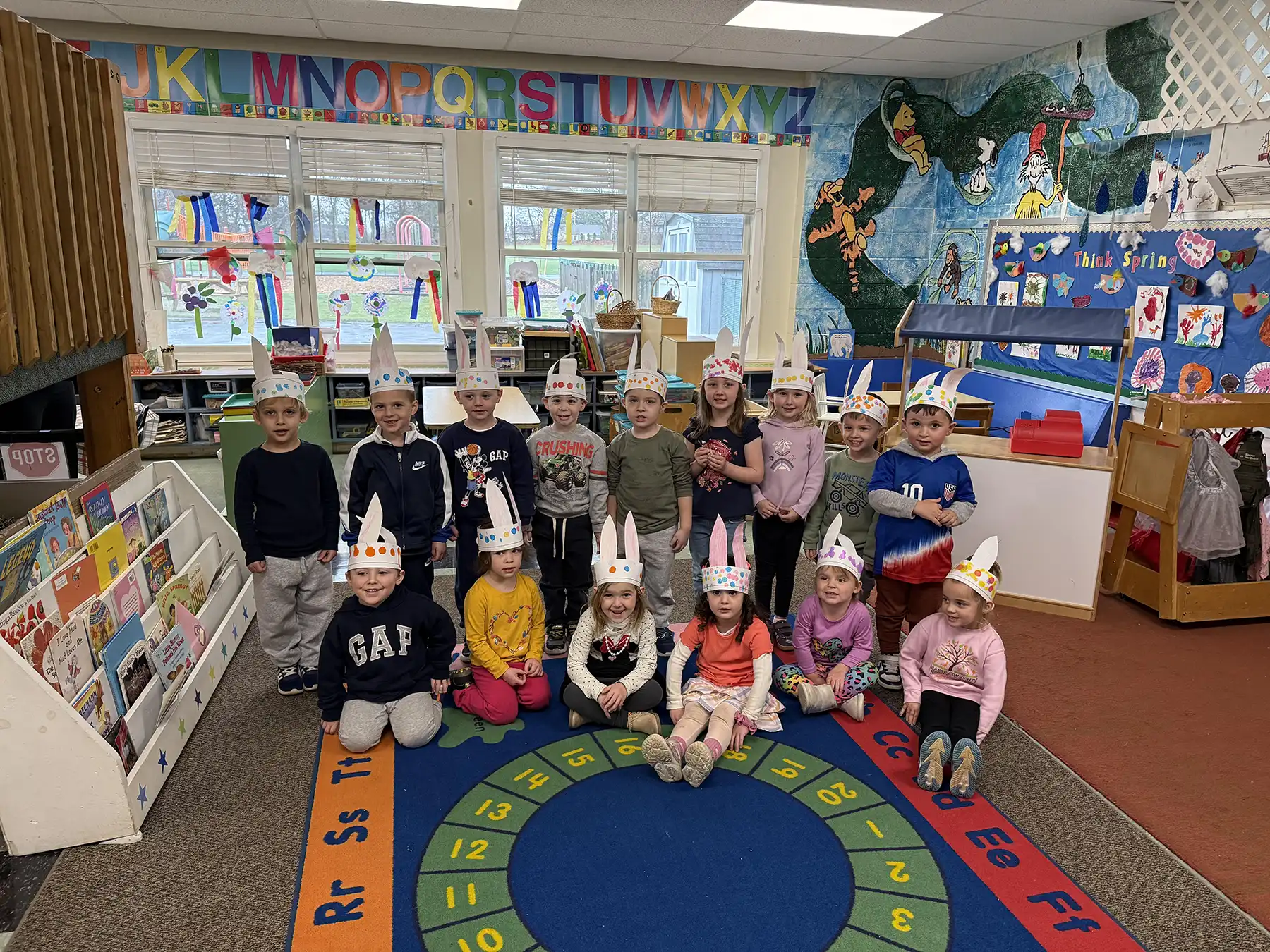 Preschoolers at Orange Community Nursery School wear handmade bunny hats while posing together in a classroom to celebrate Easter and the spring season.