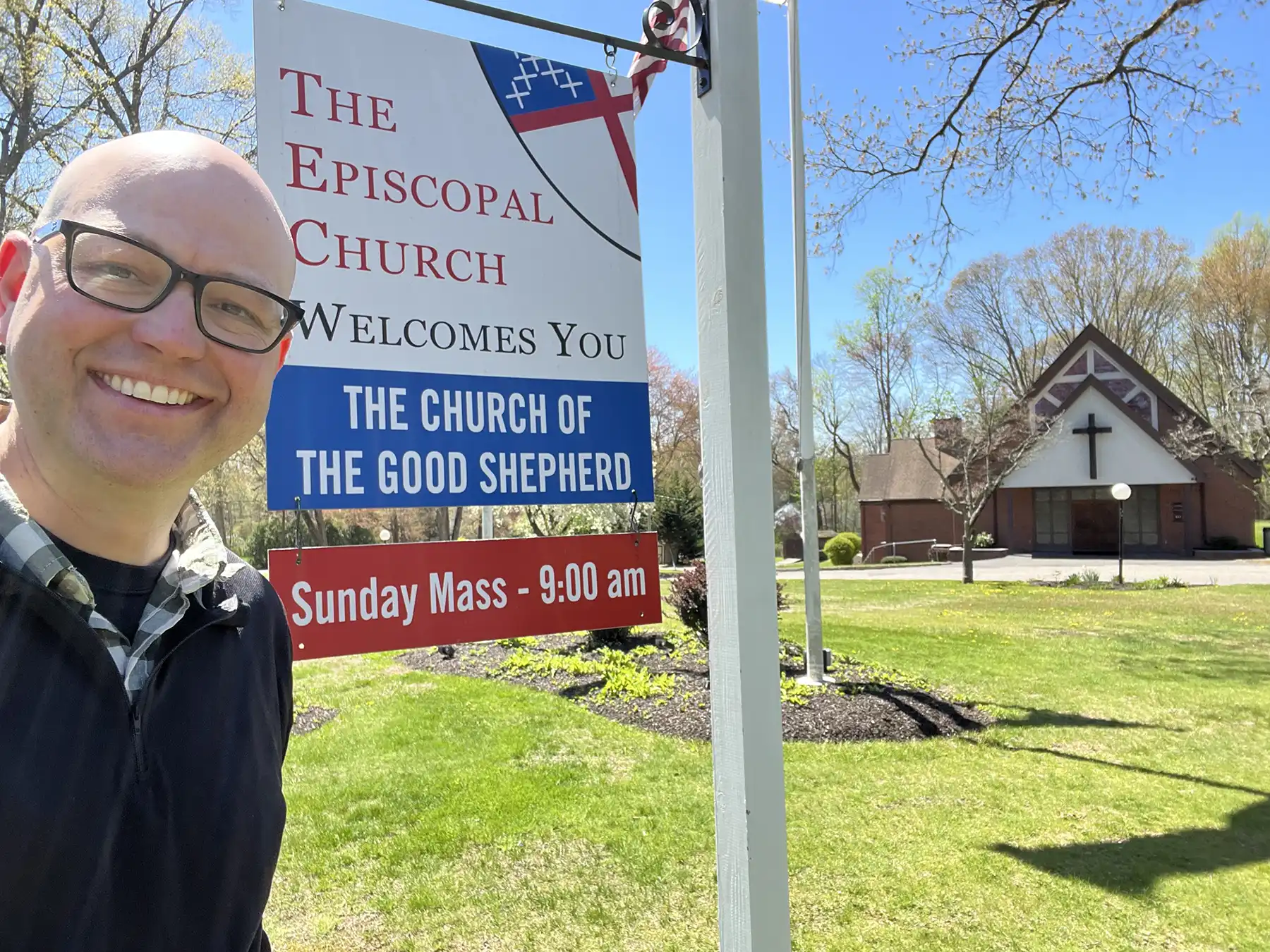 Smiling Rev. Geoff Sinibaldo taking a selfie next to The Church of the Good Shepherd sign on a sunny day with the brick church building behind.