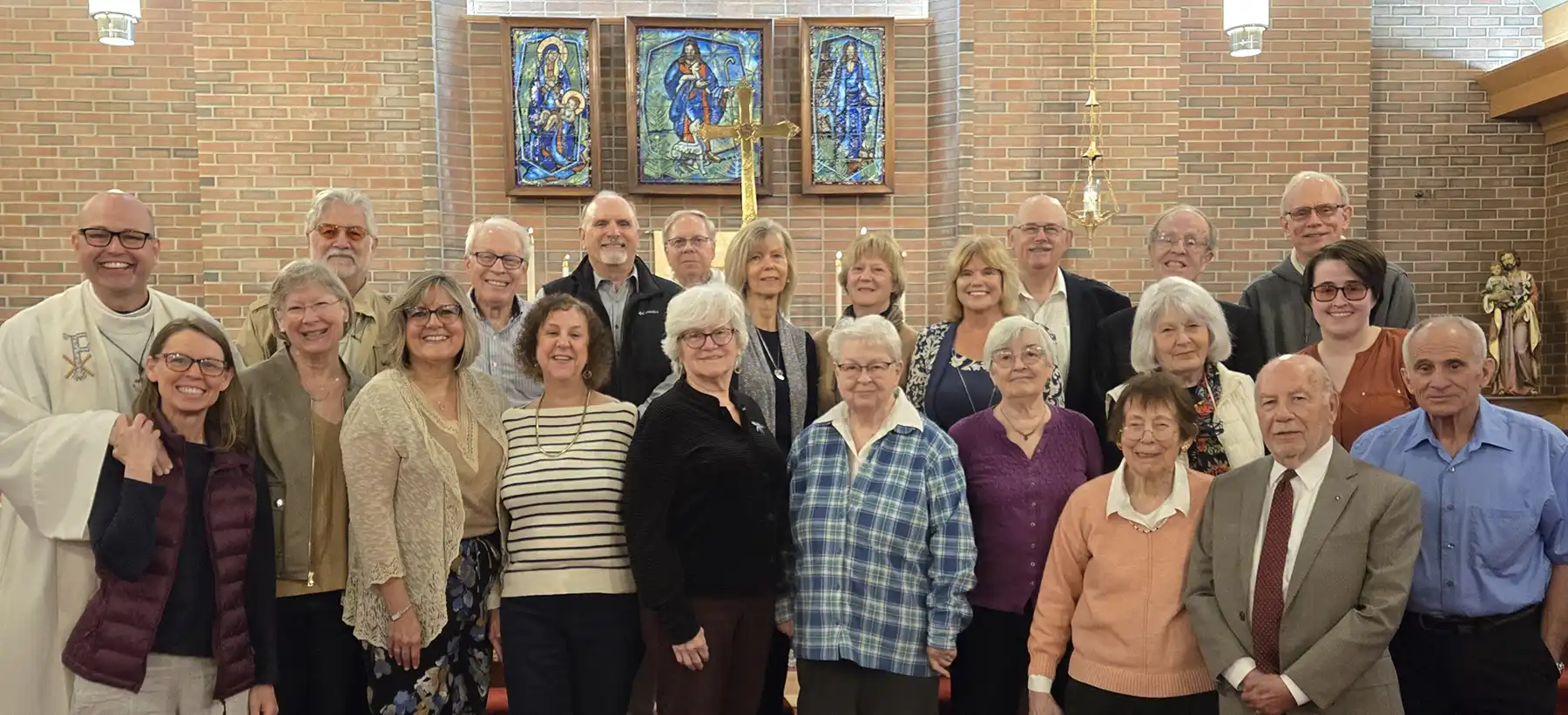 Rev. Geoff Sinibaldo poses with a diverse congregation group inside a church with brick walls, stained glass windows, and an altar cross behind them.