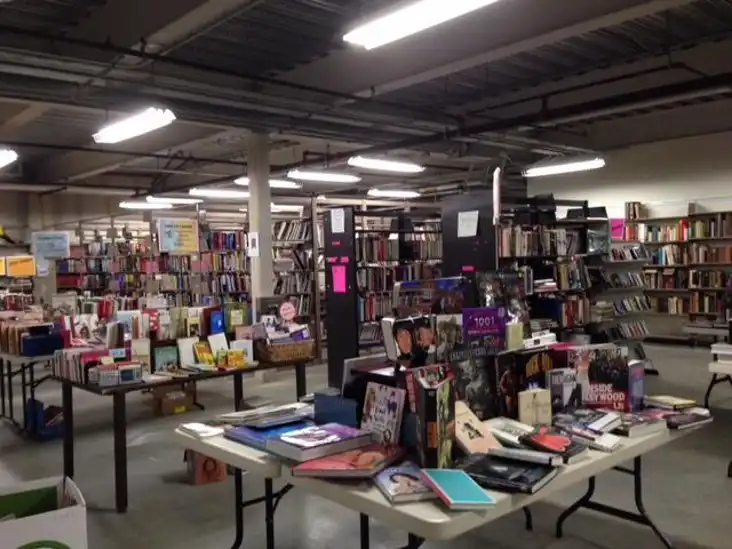 Multiple folding tables displaying rows of books in a well-lit library basement setup, with tall bookshelves packed with novels and non-fiction titles visible in the background.