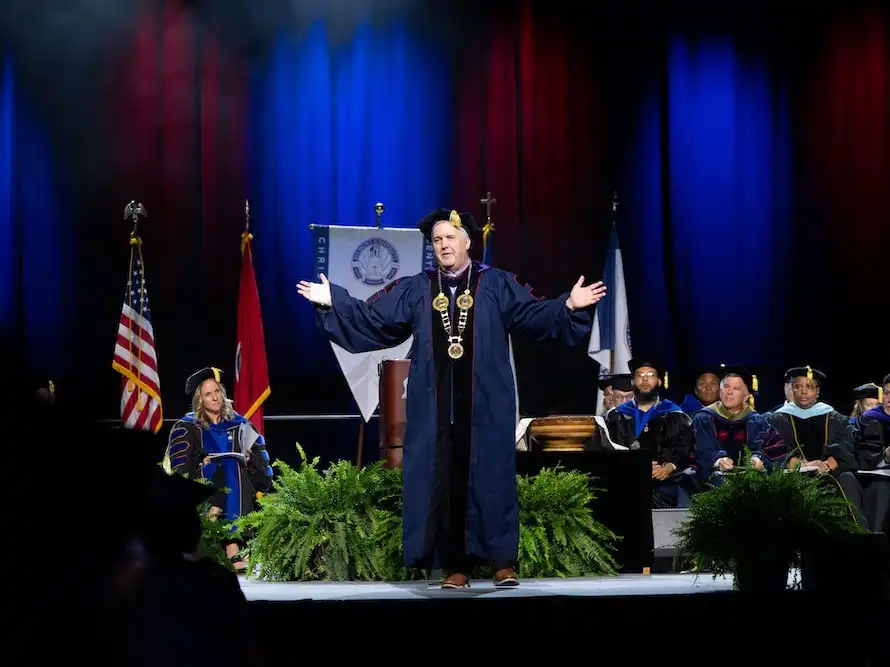 Belmont University President Dr. Greg Jones stands on a lit stage in academic regalia with arms outstretched, addressing the graduating Class of 2026.