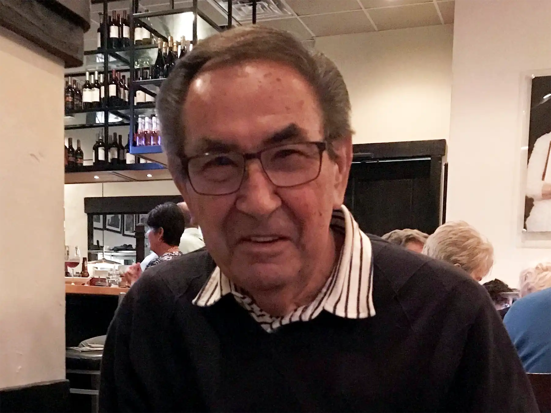 Portrait of John Peter Kowal wearing glasses and a dark sweater, smiling indoors at a restaurant with wine bottles visible in the background.