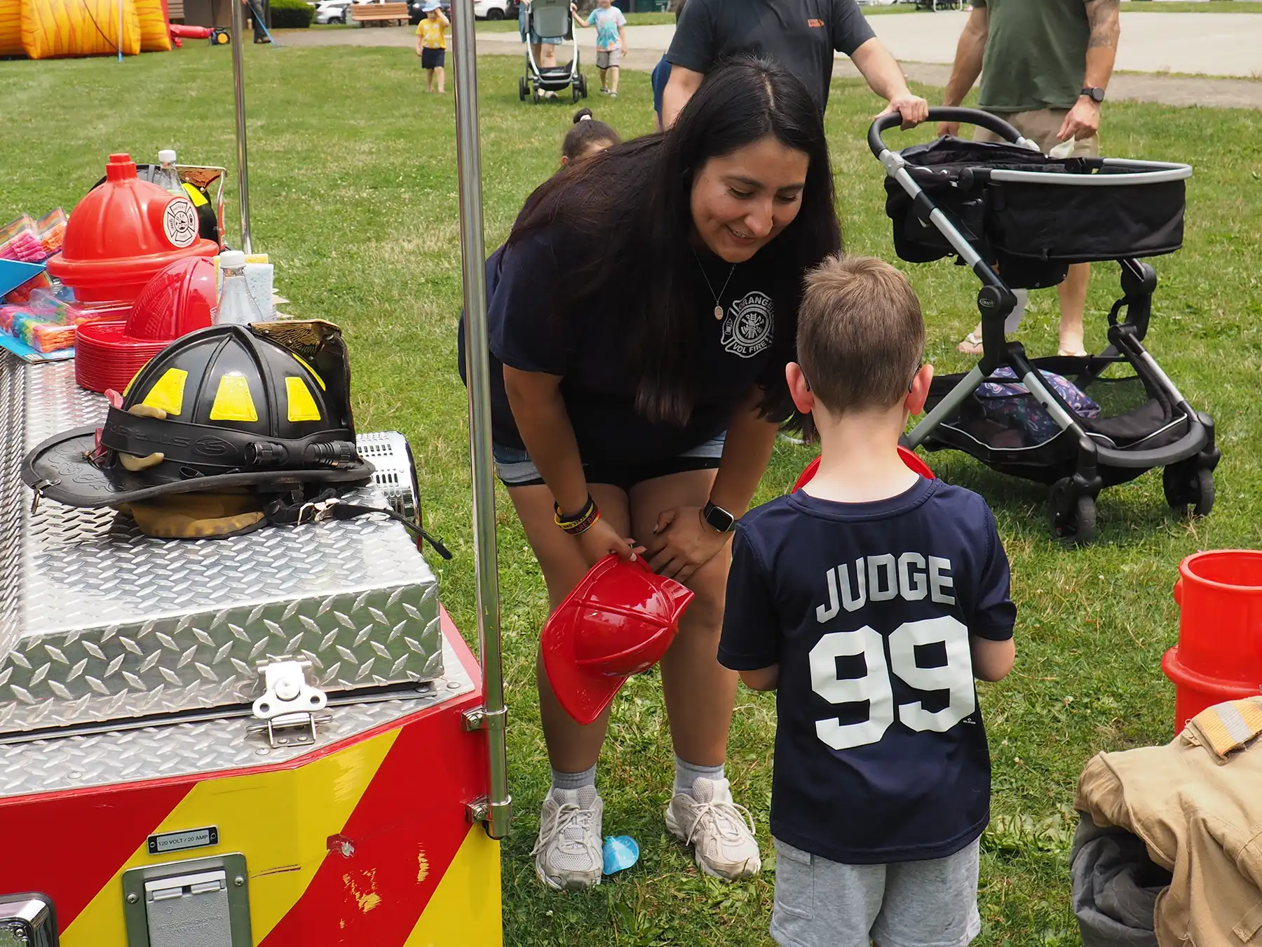Orange firefighter Wendy Camacho gives a plastic red toy fire helmet to a young boy during the town Safety Day event outdoors.