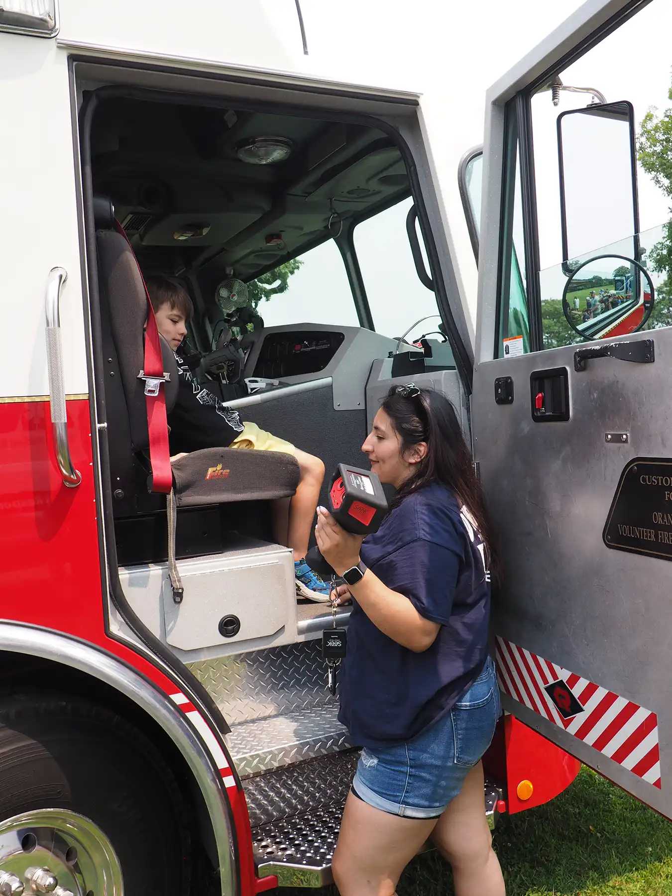 A female Orange firefighter shows a thermal imaging camera to a young boy sitting inside the cab of a red fire truck.