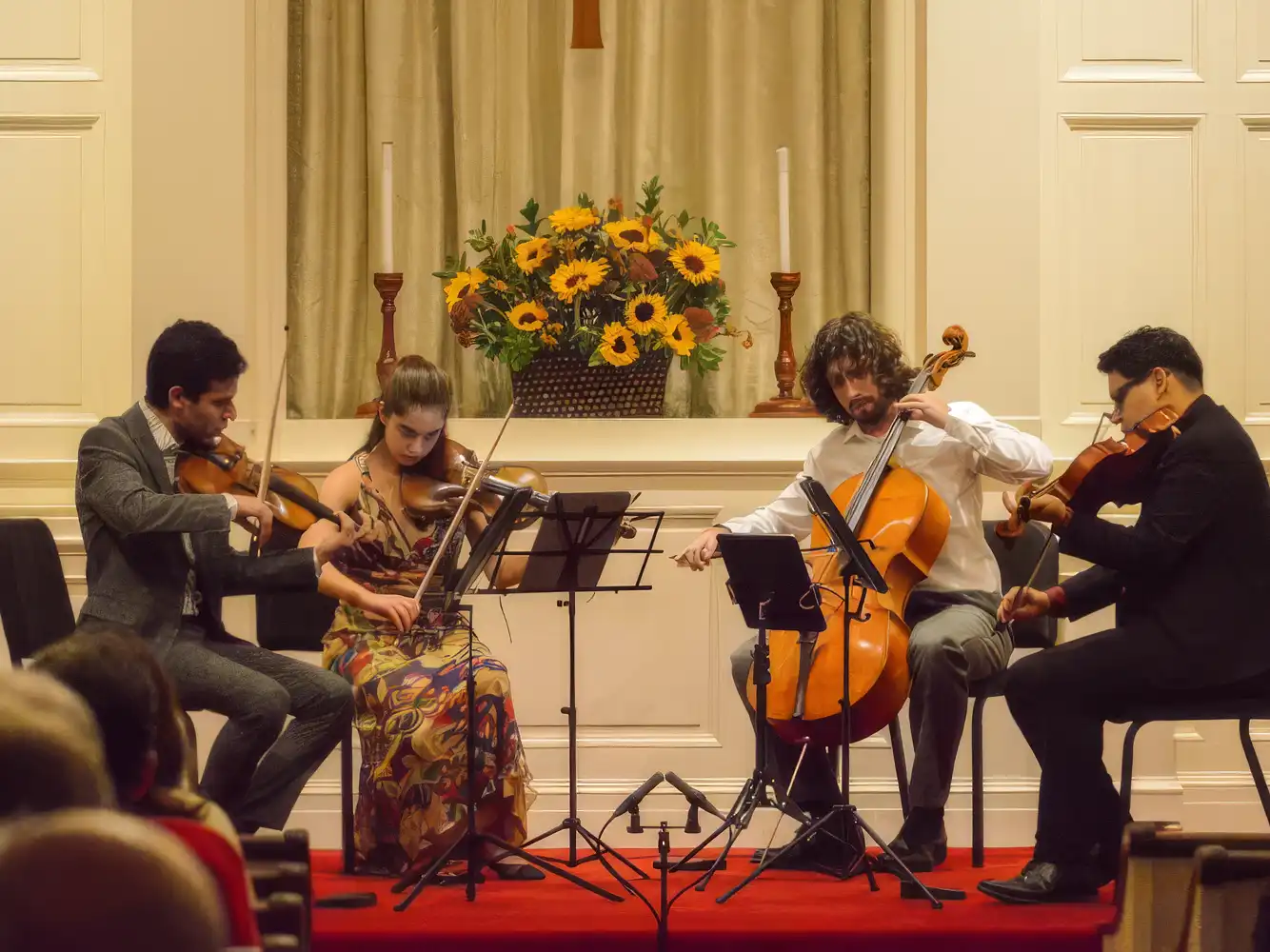 A classical string quartet with three violinists and a cellist performs live on stage in front of an audience with sunflowers in the background.