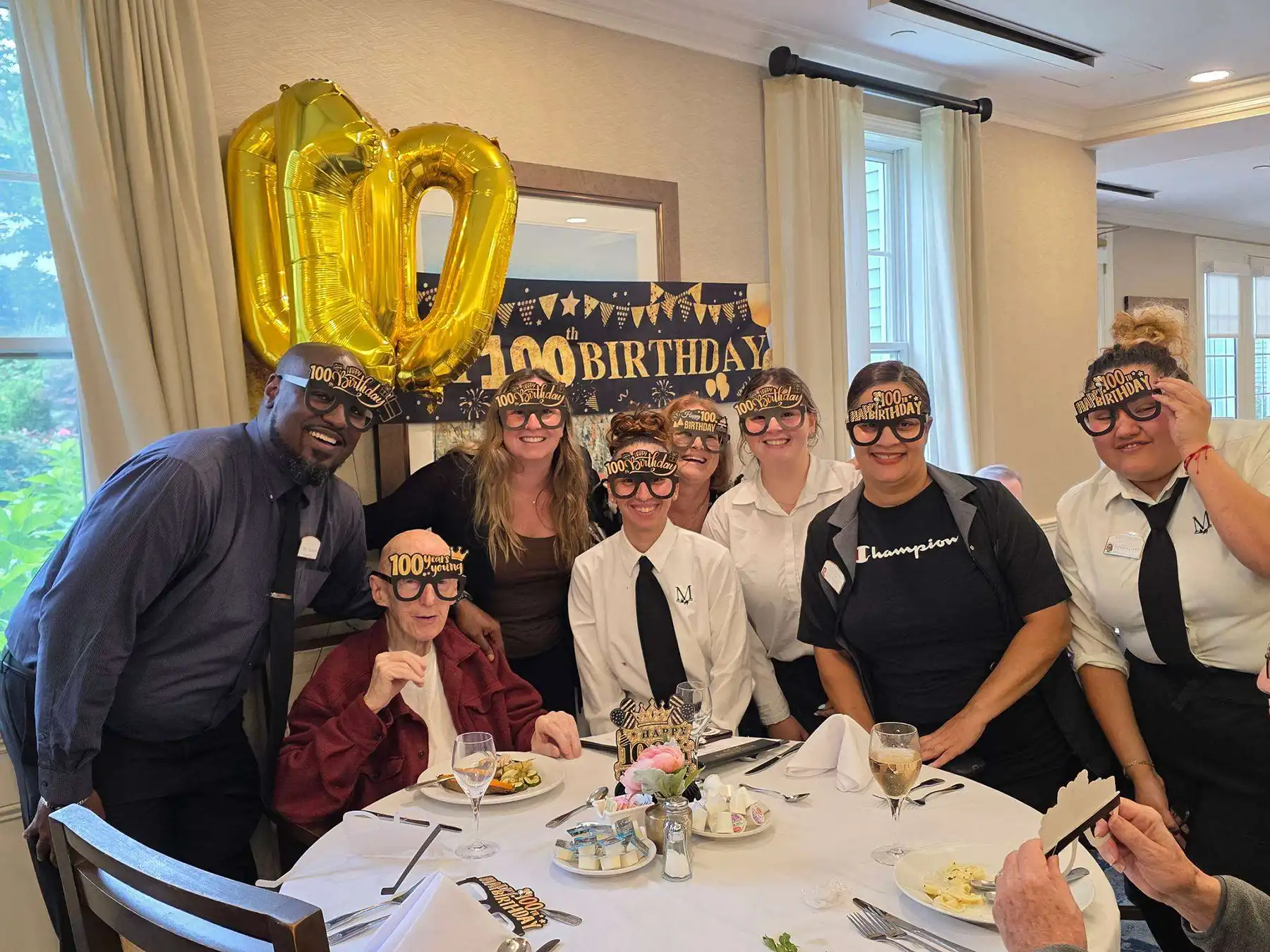 Alt Text: Maplewood at Orange staff and residents wearing novelty party glasses gather around Frank Broderick to celebrate his 100th birthday.