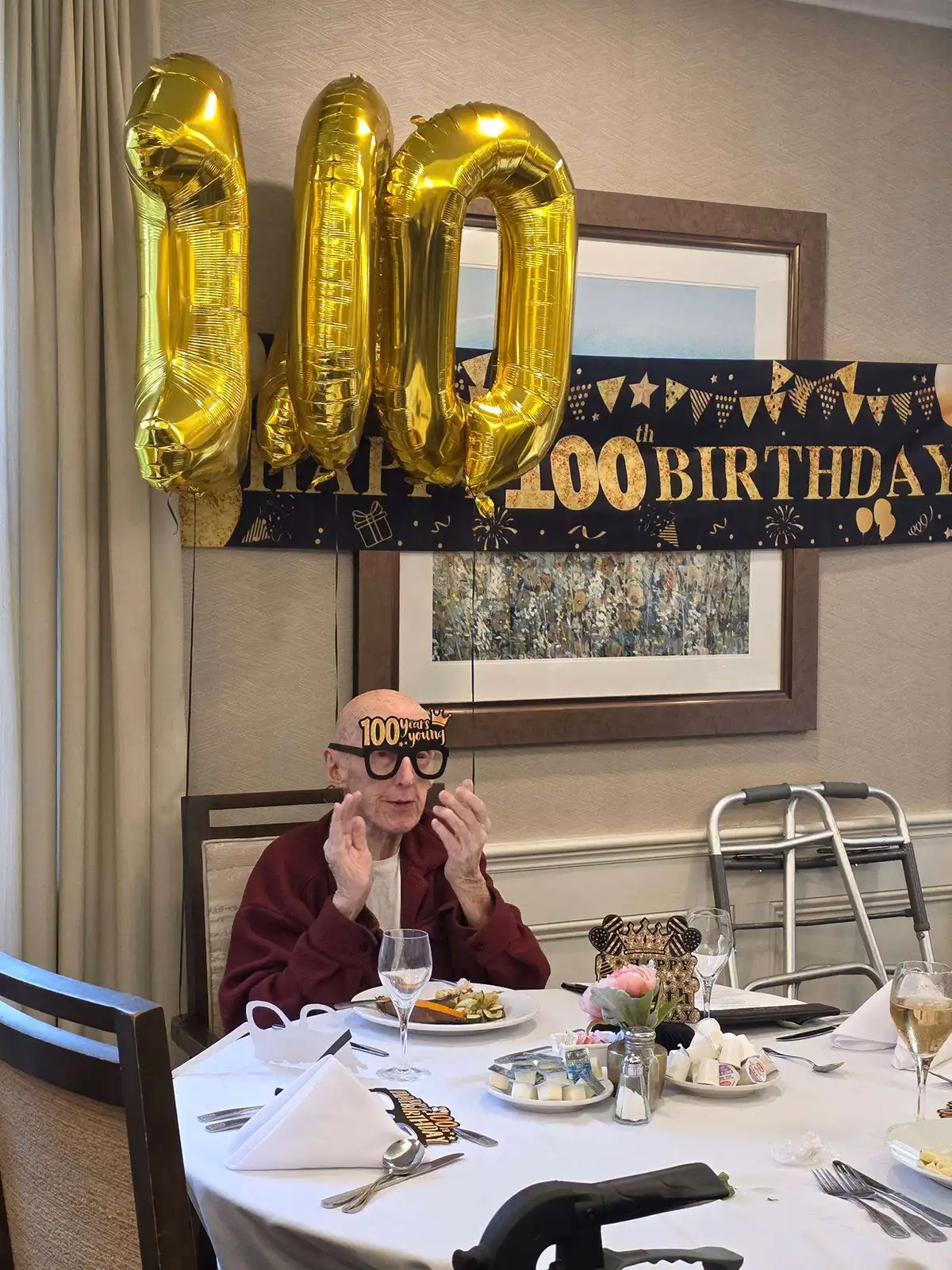 Alt Text: Frank Broderick wears 100 years young party glasses seated under gold balloons during his 100th birthday celebration at Maplewood at Orange.