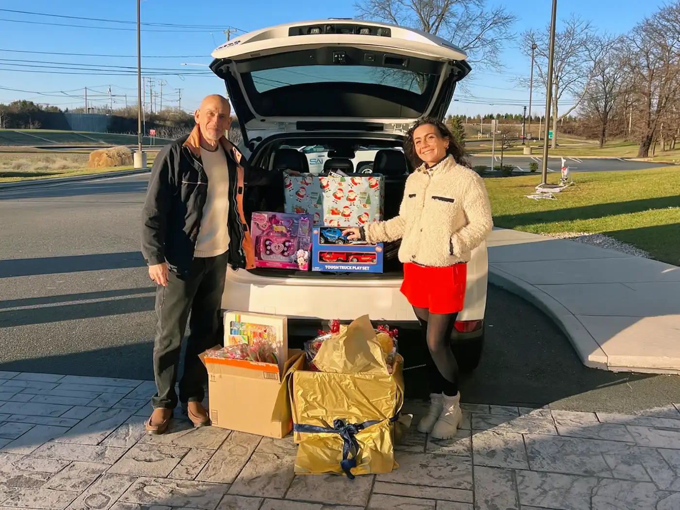 A man and woman stand by an open white SUV trunk filled with donated Christmas gifts and toys, with more gift boxes on the pavement in the foreground.