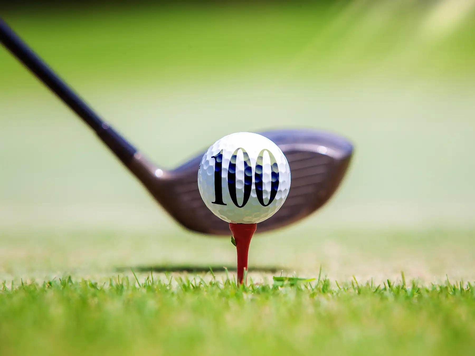 A white golf ball printed with the number 100 sits on a red tee in the grass, positioned directly in front of a driver club on a green golf course.