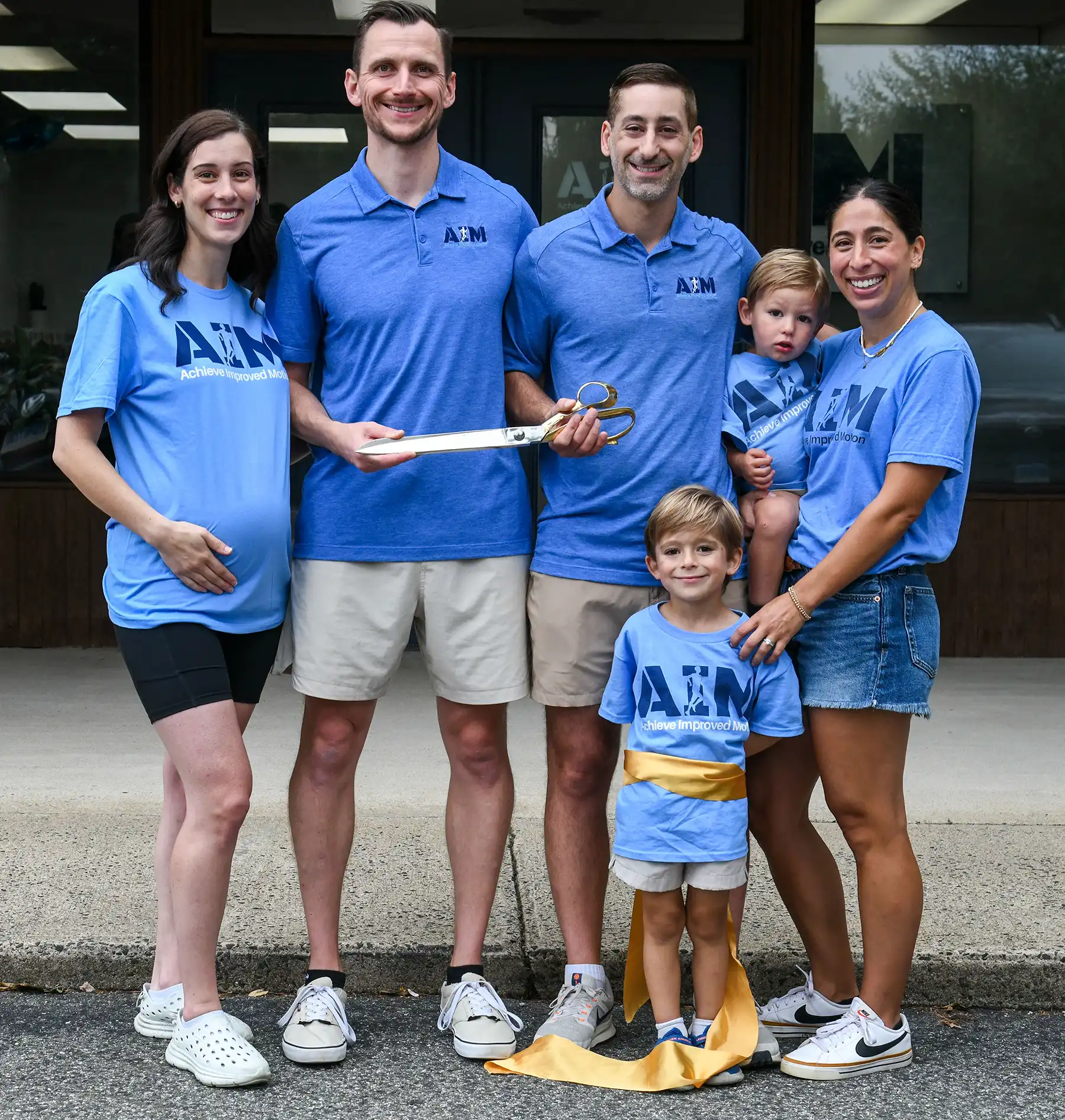Dr. Ryan Pegolo and Dr. Michael Simeoli hold giant ceremonial scissors while posing with their families in matching blue shirts outside the clinic.