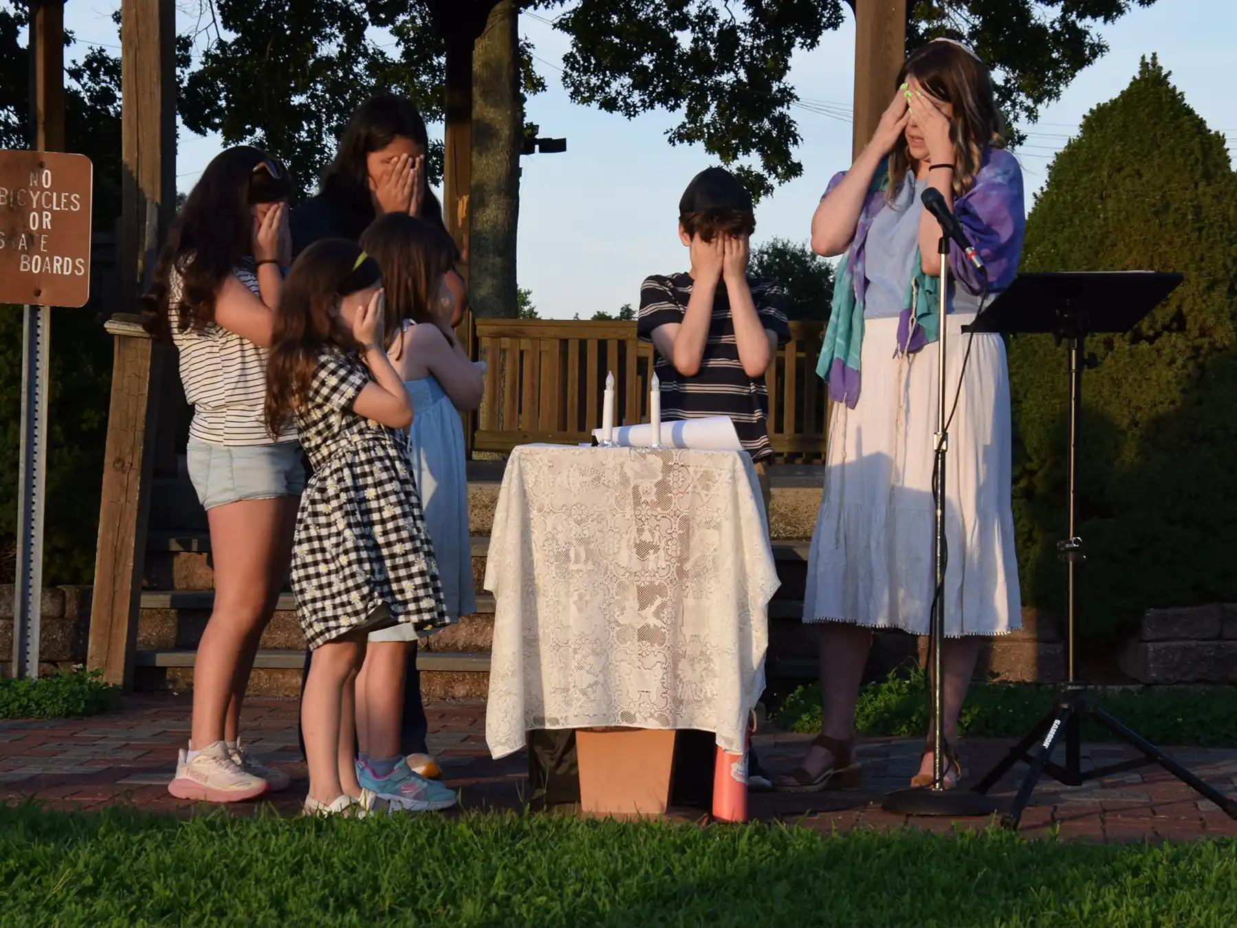 A woman and young children cover their eyes in prayer beside two lit Shabbat candles at Congregation Or Shalom’s outdoor gazebo service in Orange.