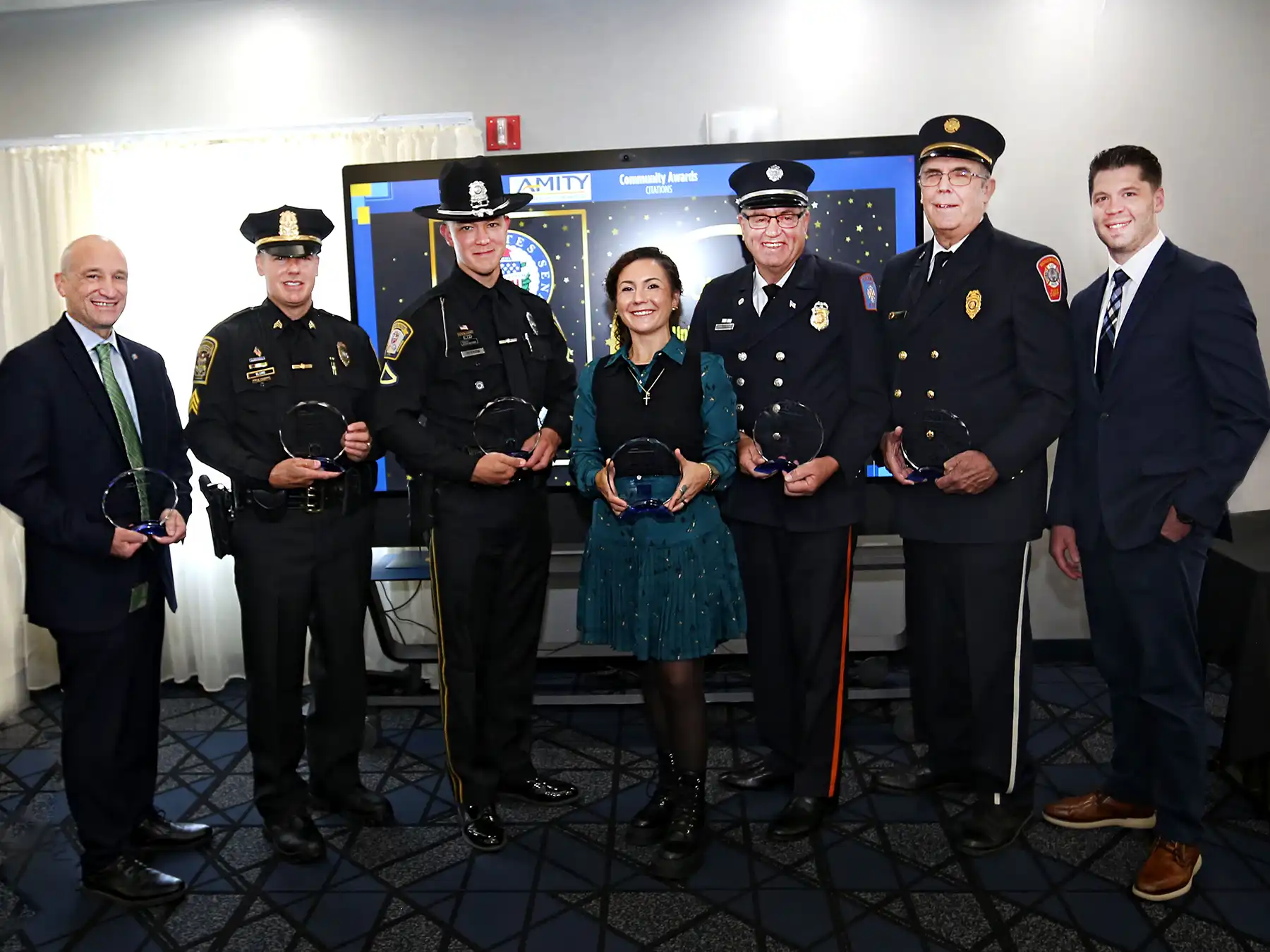 A group of police officers and firefighters in uniform pose with community leaders, holding crystal awards at the Amity Chamber event.