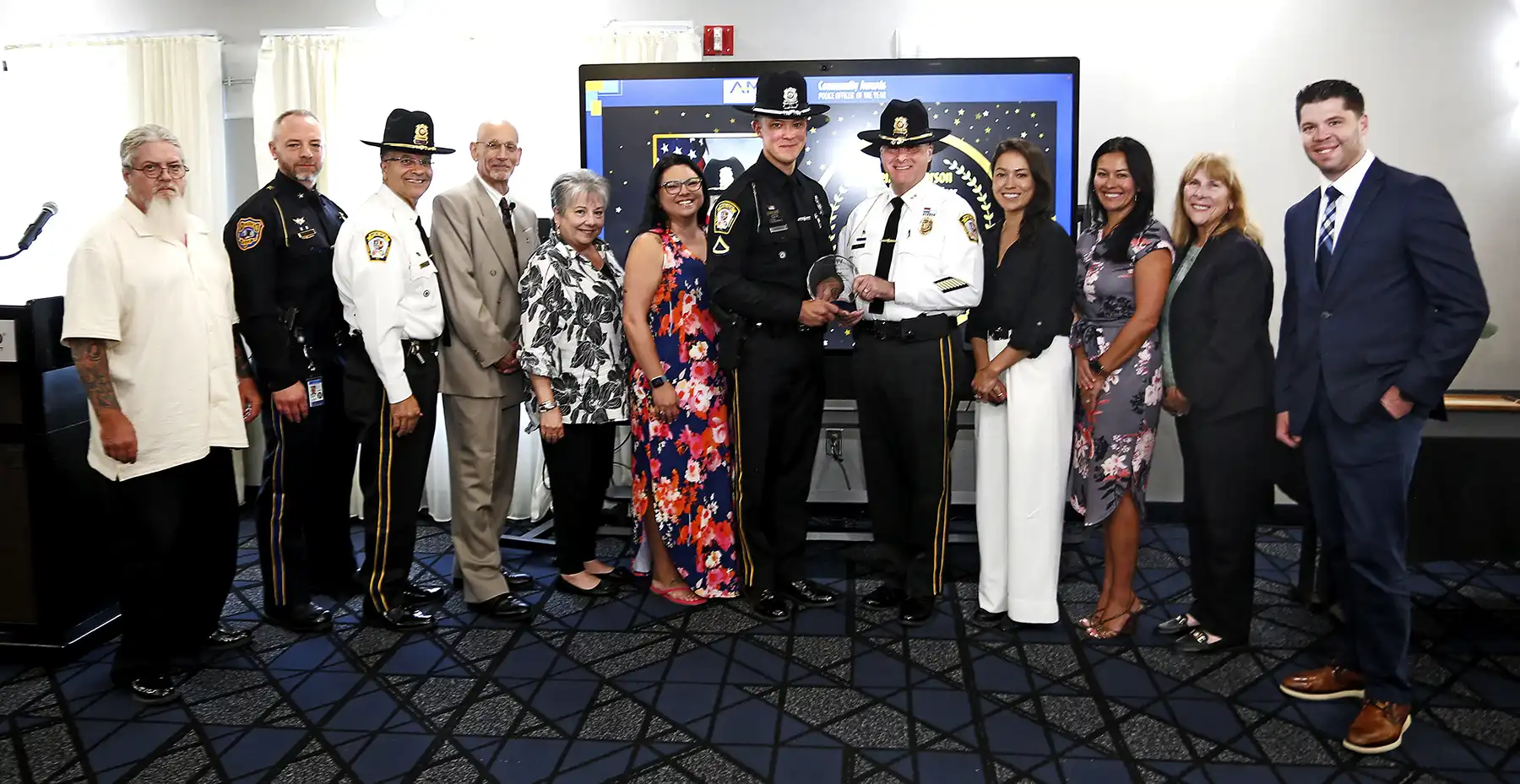 An officer in a large state trooper hat accepts an award from another officer in a white shirt, surrounded by a group of smiling civilians and officials.