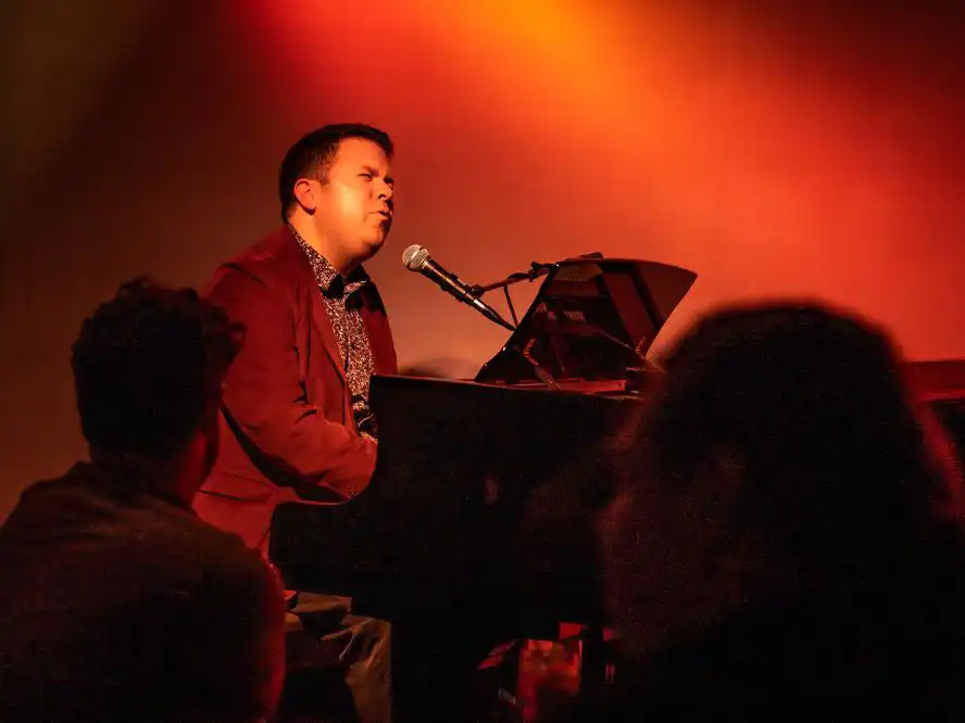 Pianist and vocalist Jay Daniels singing into a microphone while playing a grand piano under warm red stage lighting before an audience.