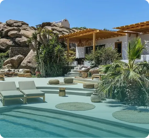 Modern outdoor patio with lounge chairs, woven cushions, palm trees, and stone steps leading to a pool and rocky hill backdrop under clear blue sky.