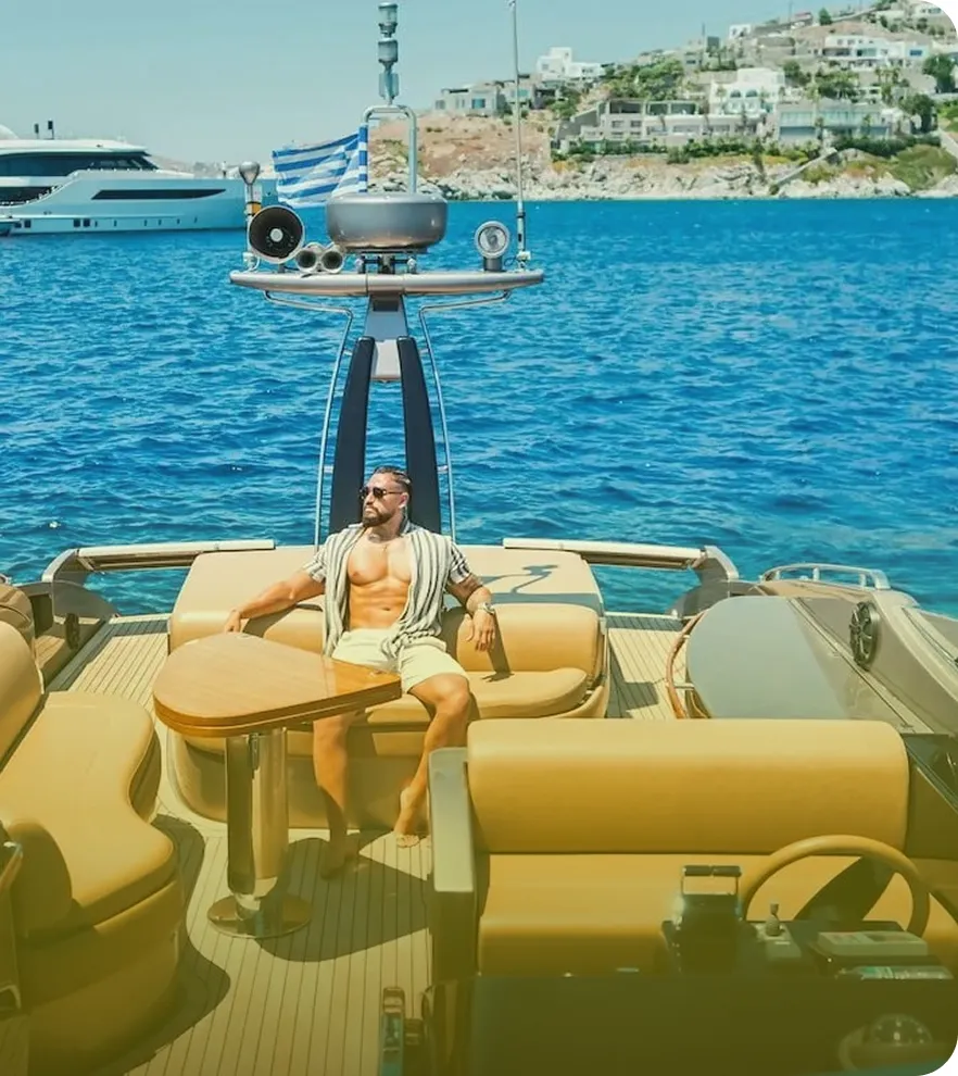 Man in sunglasses and light open shirt relaxing on the deck of a luxury yacht with blue sea and coastal buildings in the background.