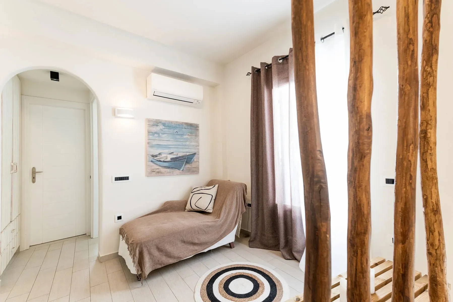 Bright room with a chaise lounge covered in a brown throw, a pillow, wall art of a boat, air conditioner, and wooden curtain rods with brown curtains.
