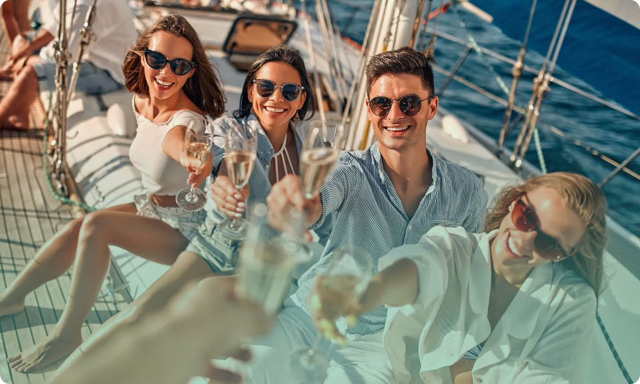 Four friends wearing sunglasses smiling and toasting with champagne glasses on a sailboat.