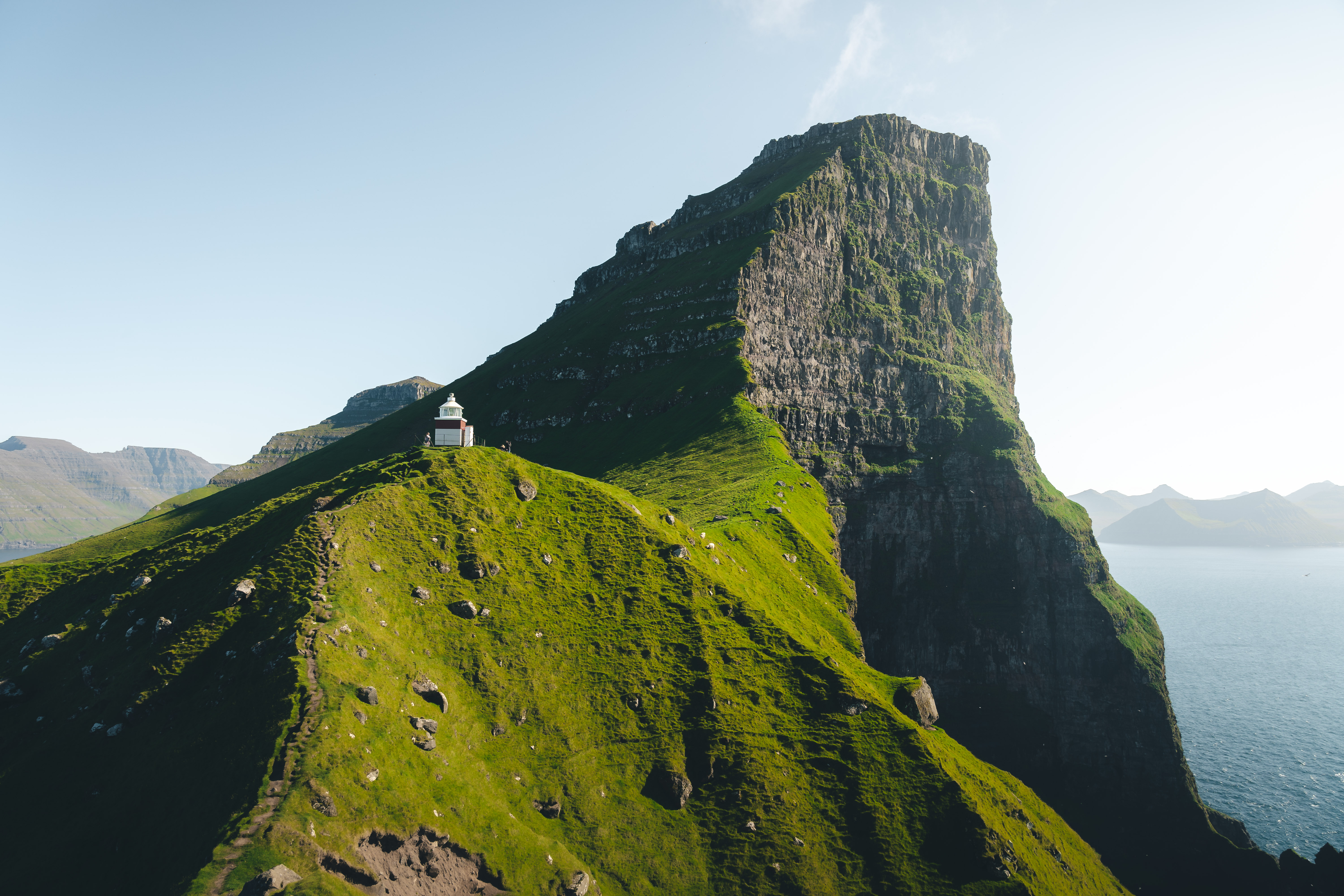Photo of a magnificent lush-green cliff controls the frame and towers over a small lighthouse in the Faroe Islands