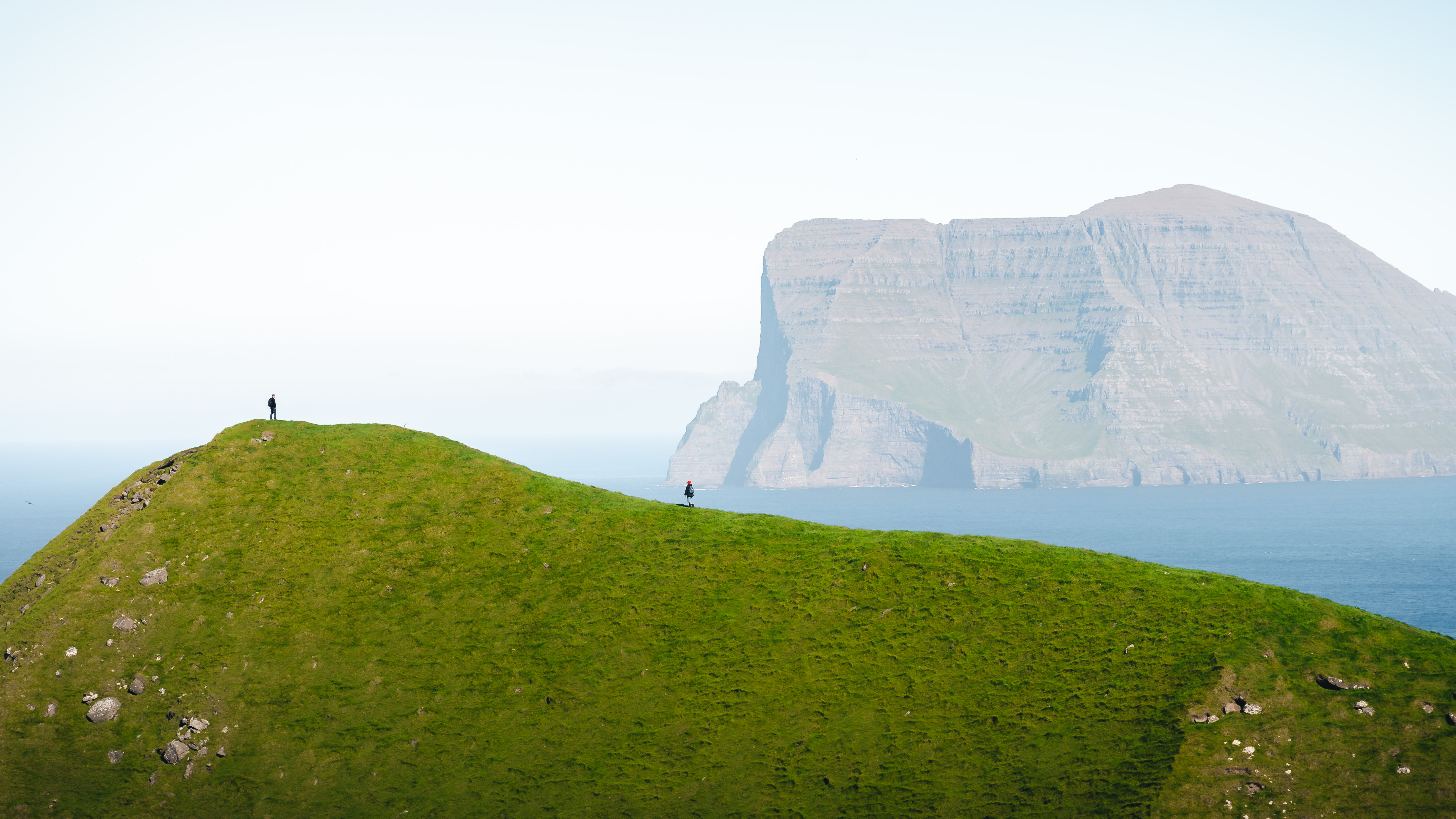 Photo of lush green hill in the foreground with two people standing on it in the distance, in the background a cliff controls the frame