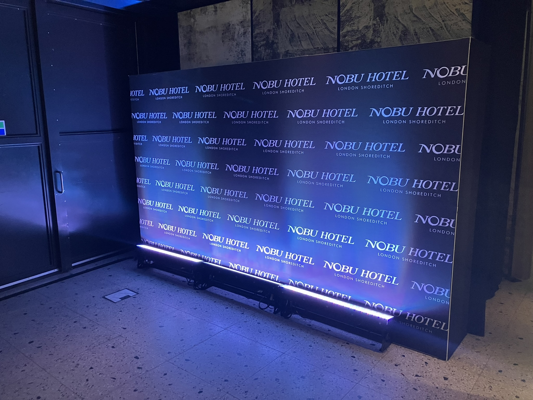 3.6 meter wide Step and Repeat Wall on hire in Shoreditch, east London - uplighting & black background with white Novikov logos on the back step and repeat wall - Product Example Photo 20