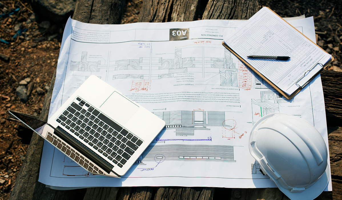 Construction plans spread across a jobsite table with a laptop, clipboard, and hard hat, illustrating how builders manage project documents across multiple tools and paper files.