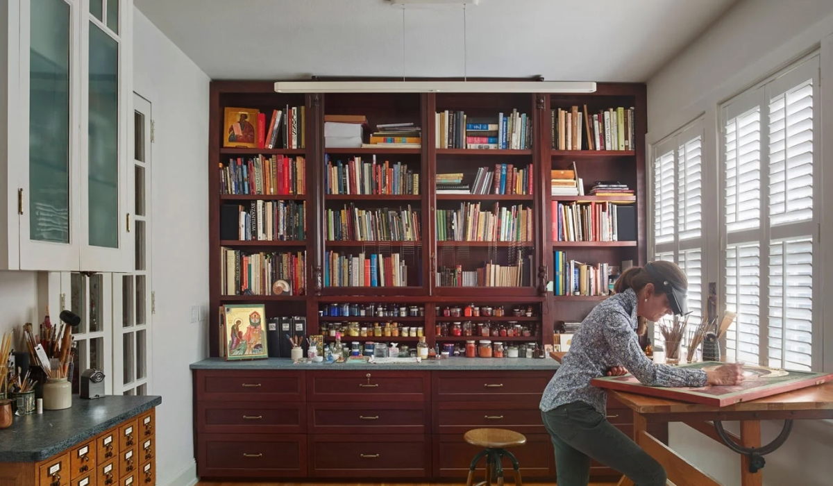 Woman working at a drafting table in a home art studio lined with built-in bookshelves and an antique card catalog.
