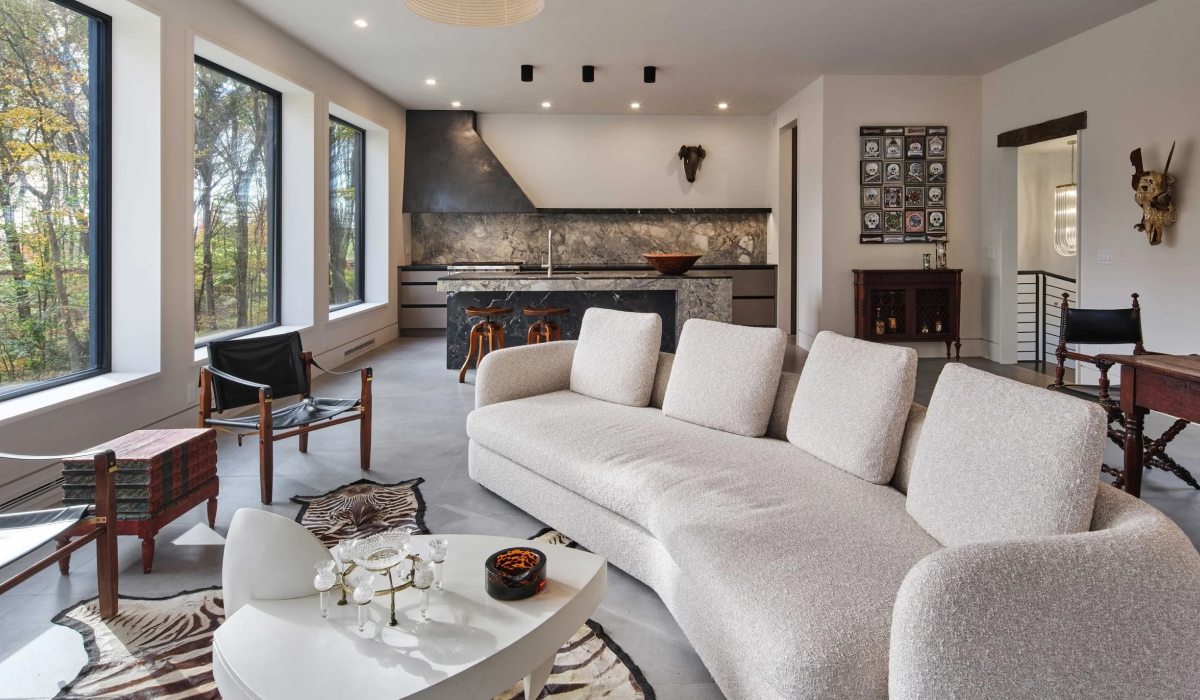 Modern living room with a curved boucle sofa, marble kitchen island, and floor-to-ceiling windows overlooking trees.