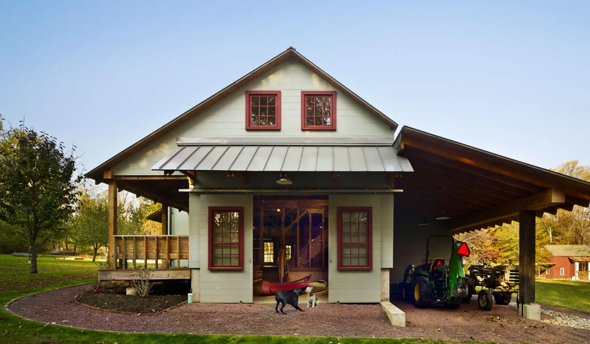 Farmhouse-style barn with red-trimmed windows, a covered porch, and an attached open-air garage housing tractors, with two dogs at the entrance.