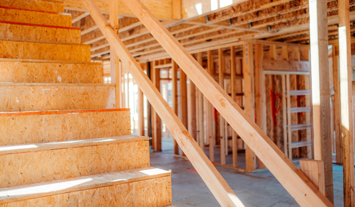 Interior view of a home under construction, showing unfinished wooden stairs and exposed wall framing lit by natural light