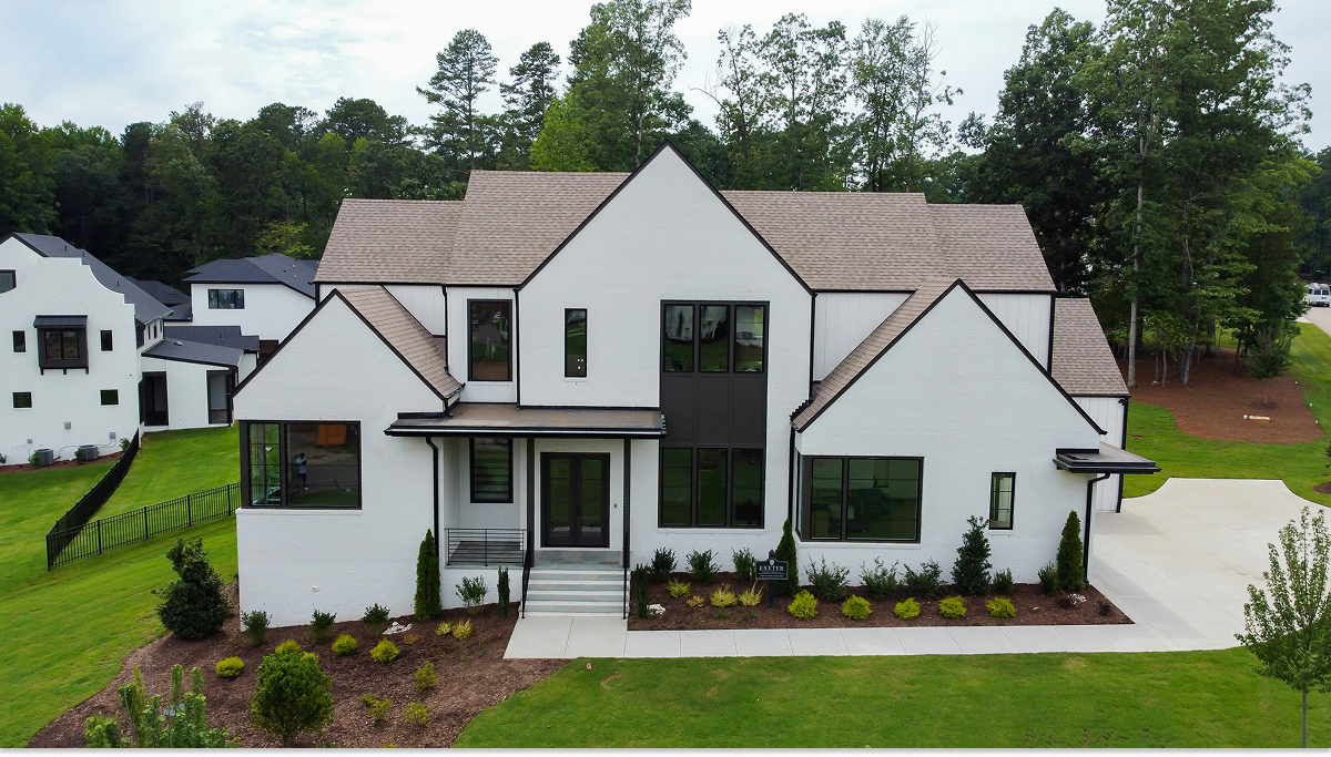 Aerial view of a white two-story modern farmhouse-style home with black trim, a shingled roof, and landscaped front yard, with additional homes visible in the background.