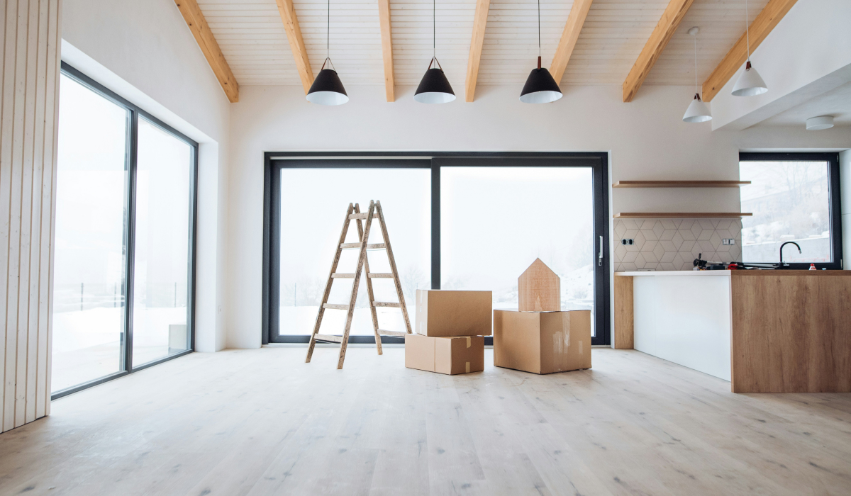 Empty, newly finished home interior with light wood beam ceilings, large windows, and moving boxes and a ladder near the kitchen area.