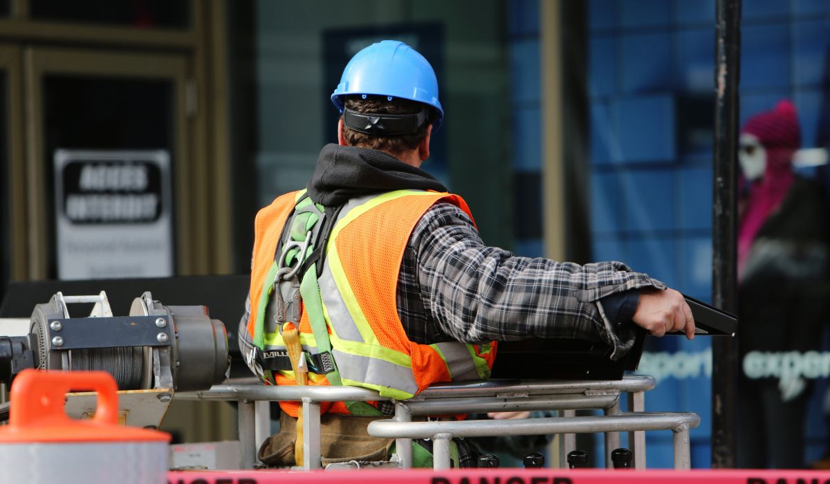 Construction worker in a blue hard hat and orange safety vest, seen from behind, operating equipment on an elevated platform at a job site.