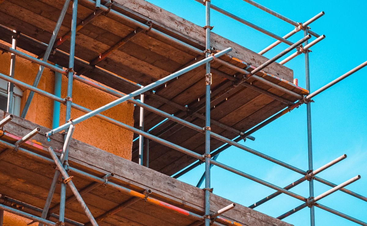 Low-angle view of wooden scaffolding platforms and metal scaffold framing against a stucco building exterior and blue sky.