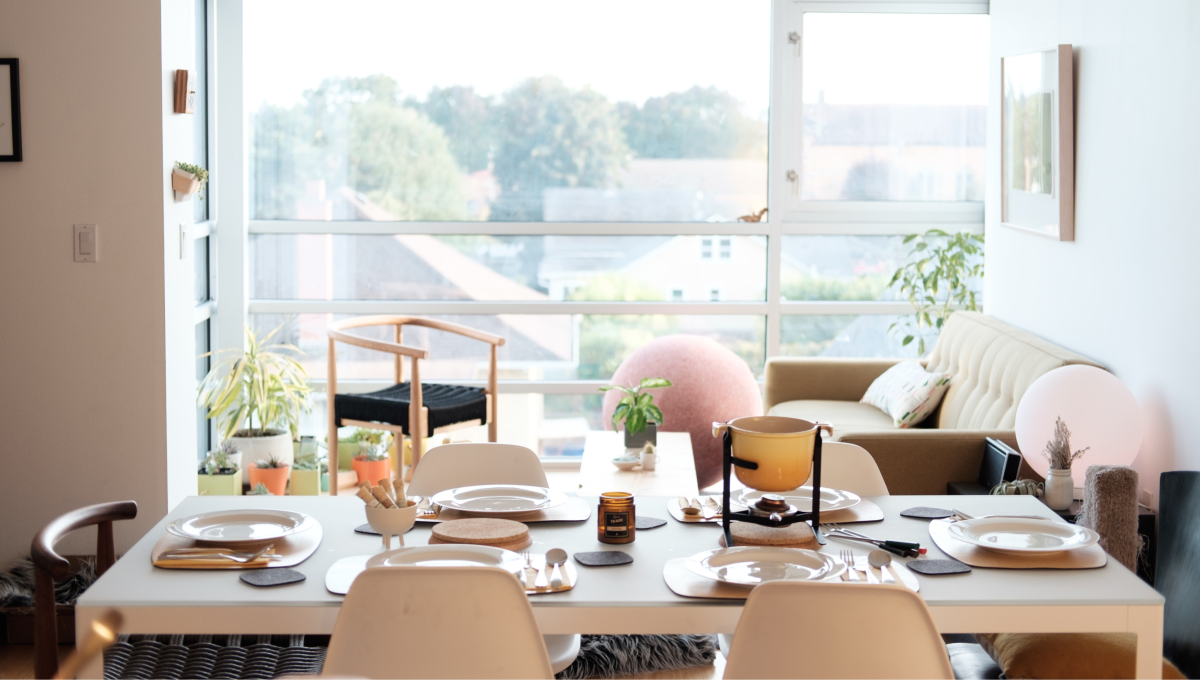 Dining table set for a meal in front of large floor-to-ceiling windows overlooking trees and rooftops.