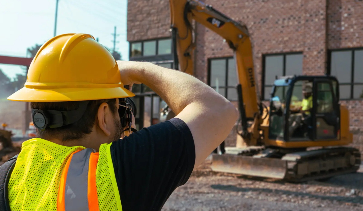 Construction worker in a hard hat and safety vest looking out at an excavator operating on a job site.
