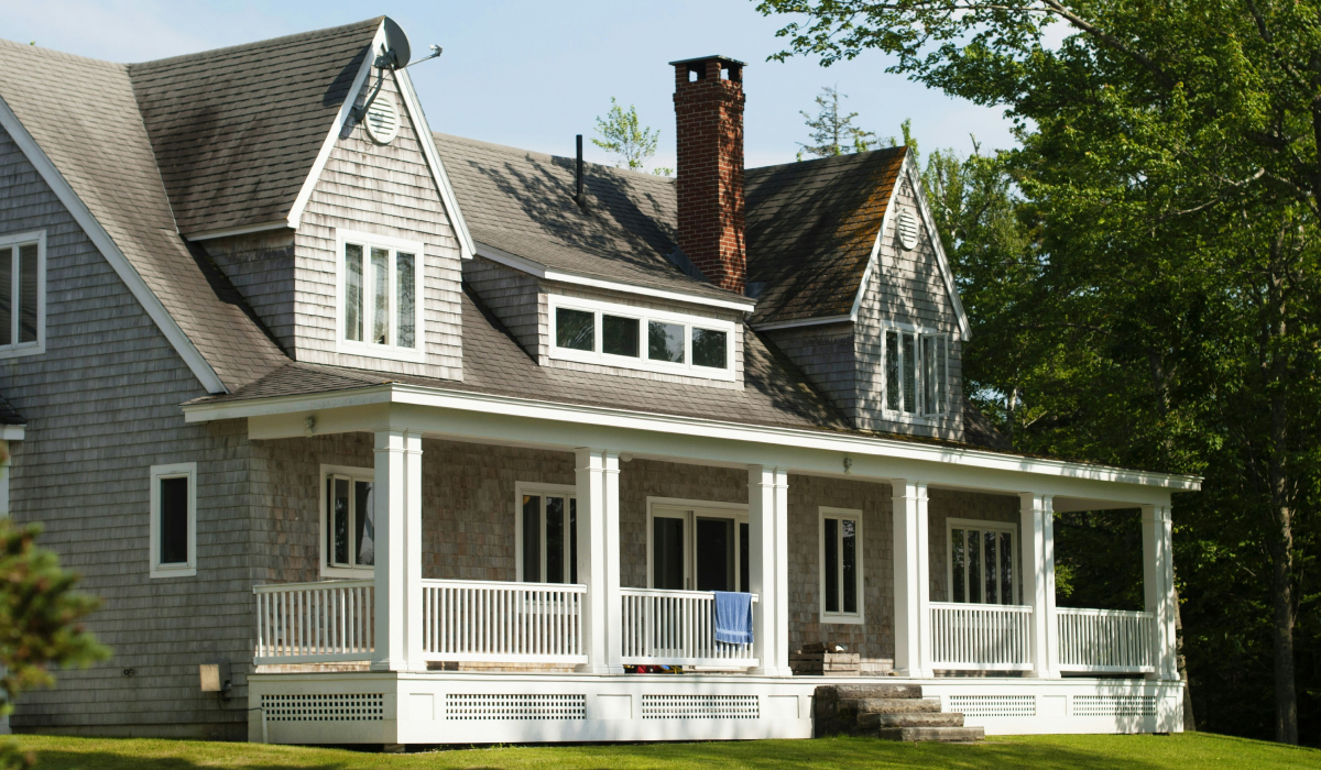 Gray shingle-style home with wraparound white porch, dormer windows, and a brick chimney