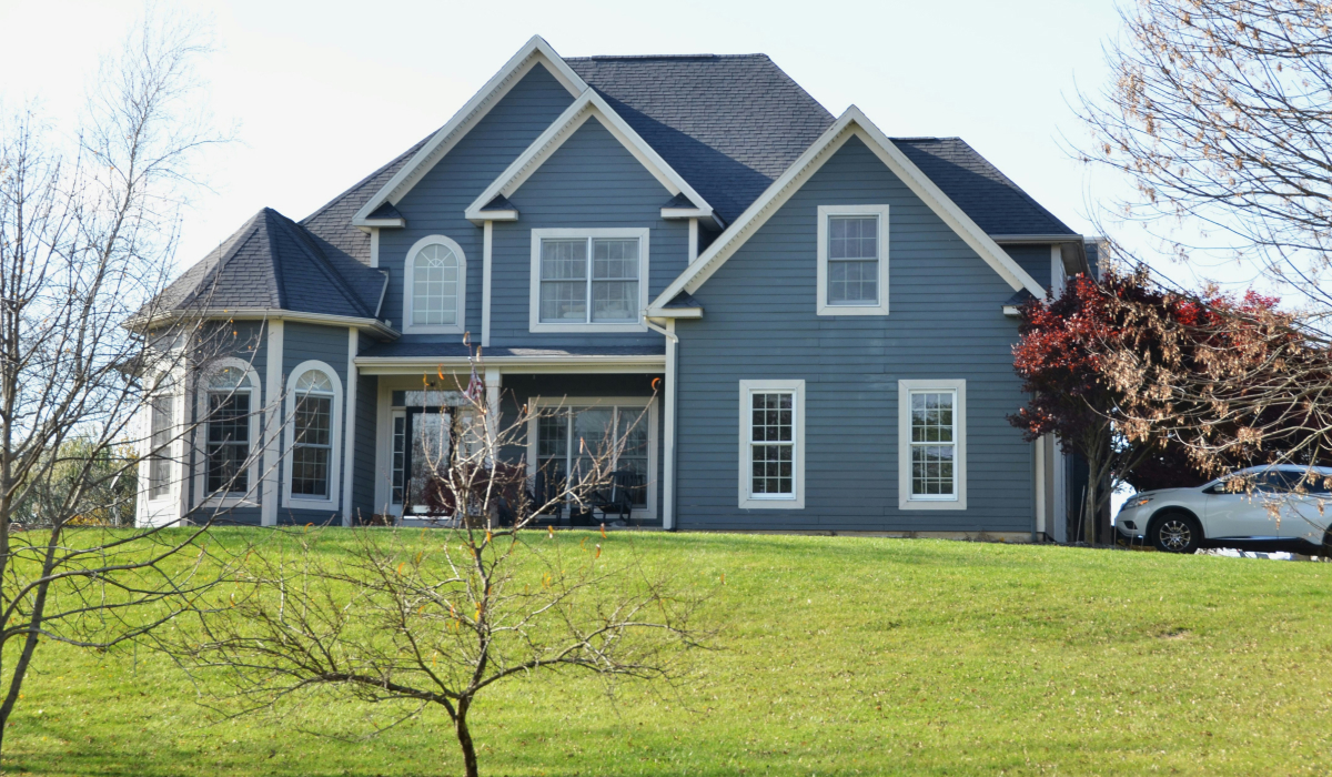 Two-story blue home with white trim and a front porch, set on a grassy hill.