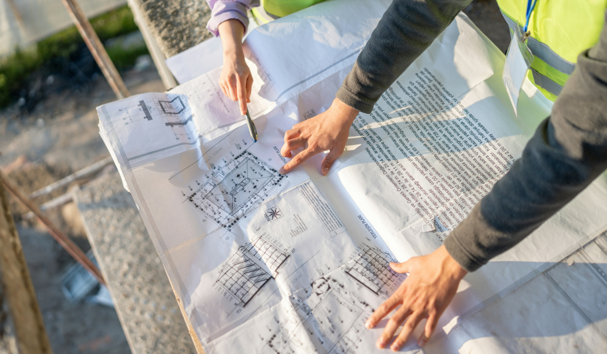Two people in work clothes review printed construction blueprints spread on a surface at a job site, one pointing at a floor plan with a pen.