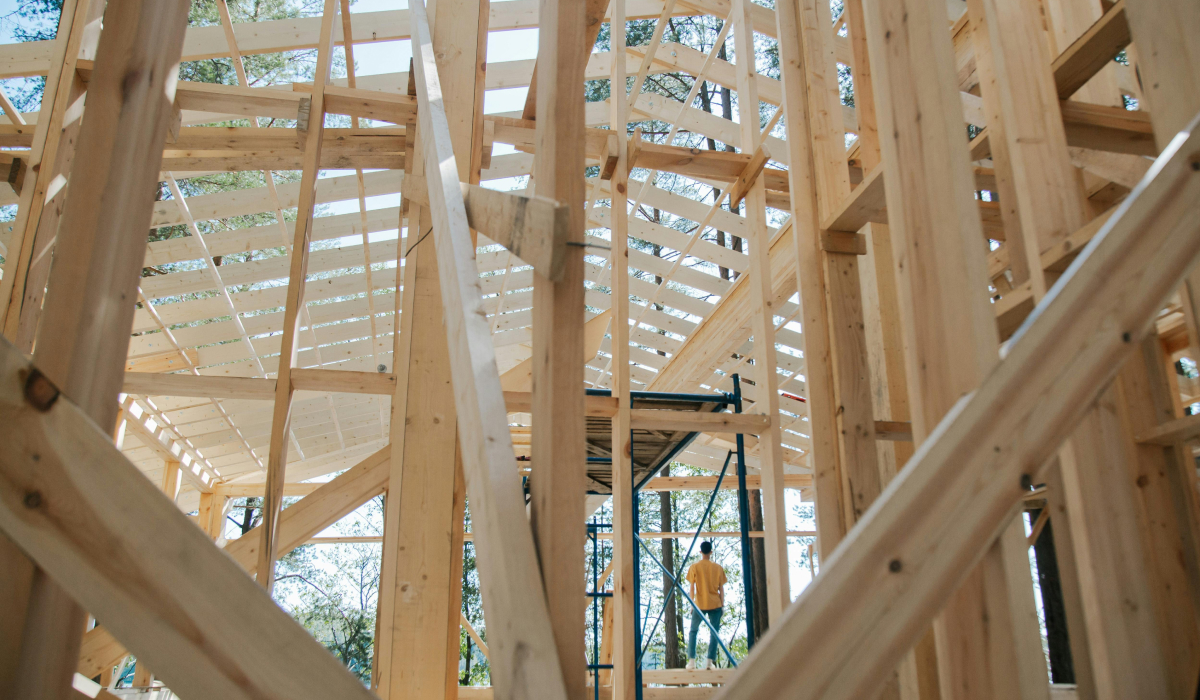 Wooden roof and wall framing of a home under construction, with a worker in a yellow shirt standing on scaffolding in the background.