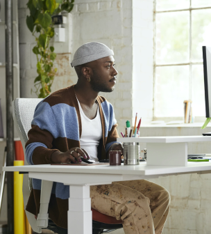 Man wearing a beanie and a striped cardigan working at a desk with a computer in a bright room with a large window.