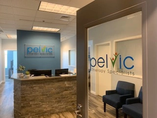 Reception area of Pelvic Therapy Specialists with a stone front desk, office chairs, and the clinic's logo on a glass door and wall.