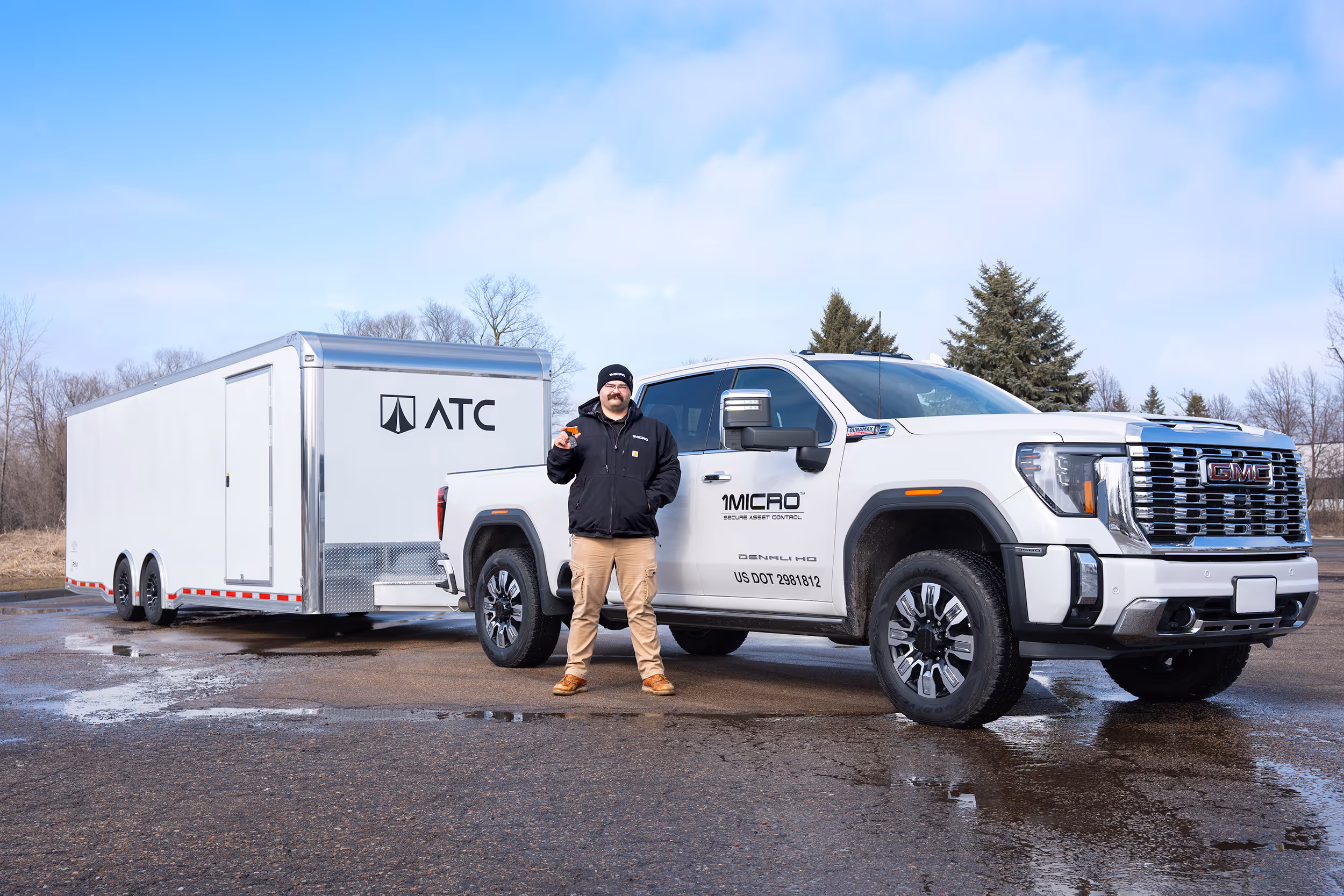 A 1Micro delivery driver wearing a black jacket and beige pants standing in front of a white GMC pickup truck towing a white trailer.
