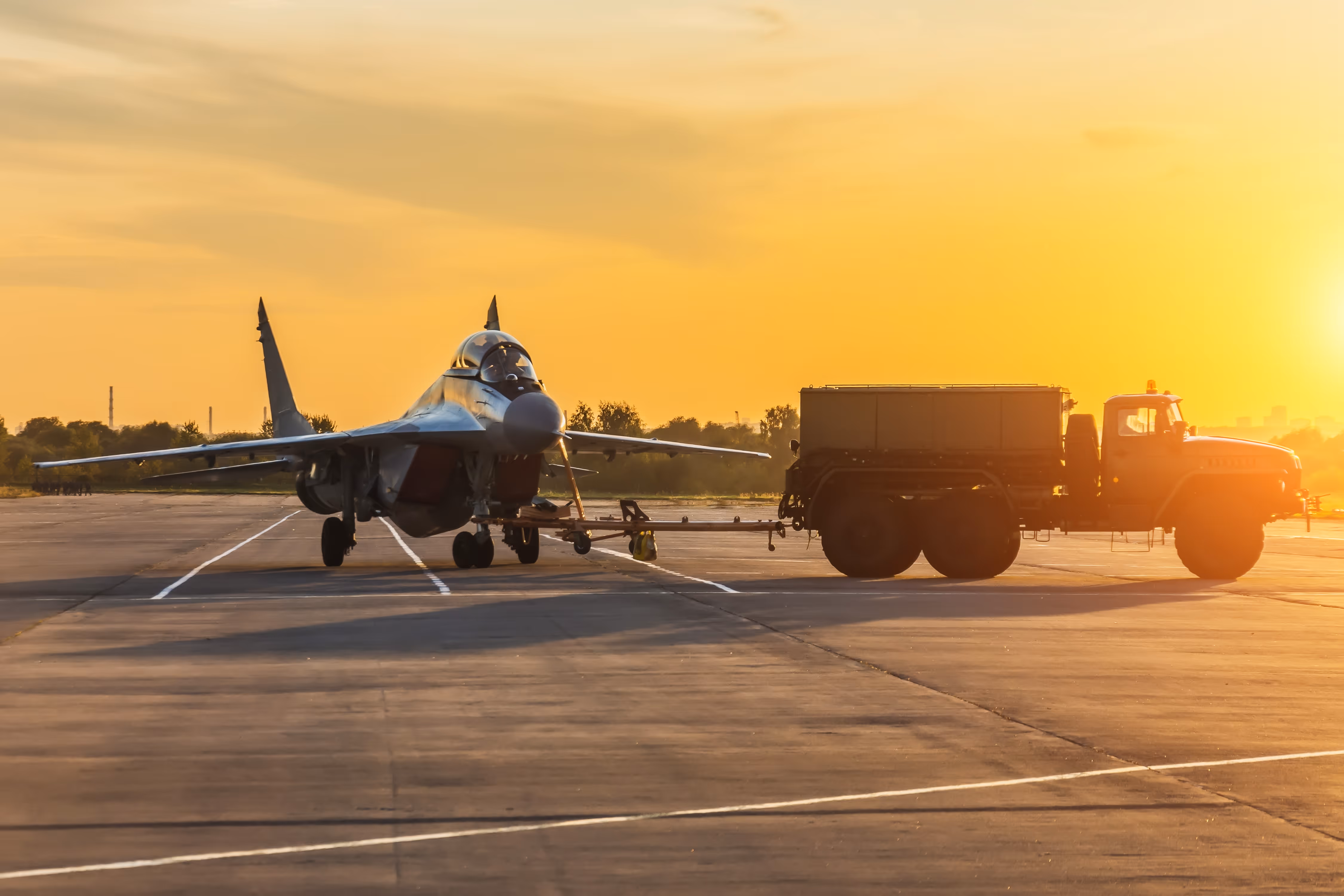 Military fighter jet being towed by a large truck on an airstrip at sunset.
