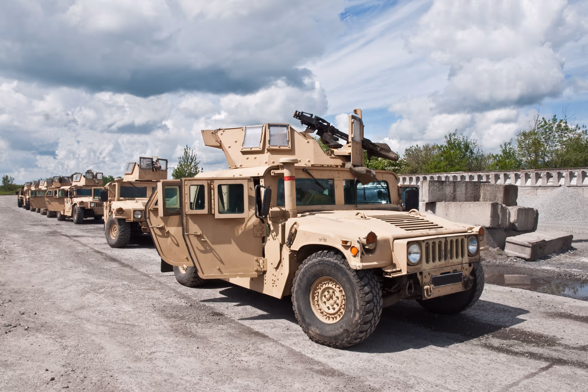 Line of beige military Humvee vehicles parked on a gravel road under a cloudy sky.