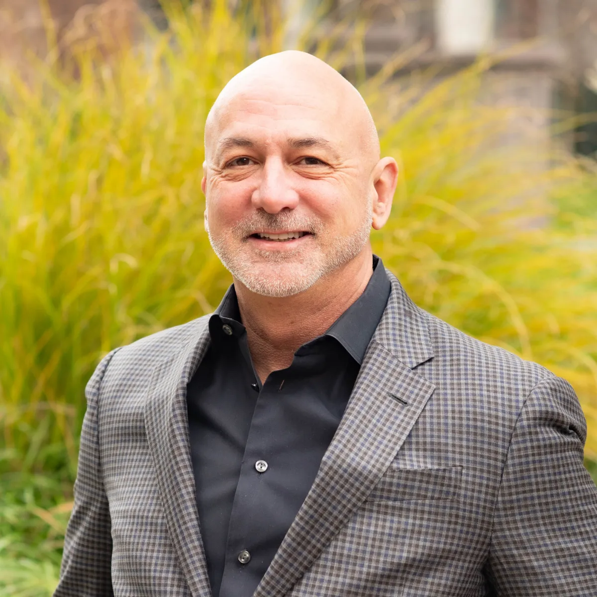 Smiling bald man with trimmed beard wearing a checked blazer and black shirt outdoors.