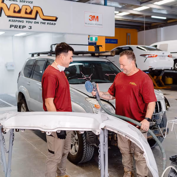 Two men in red shirts and tan pants stand in an auto repair shop next to a silver SUV, one holding a sanding tool while the other watches, with a detached car bumper in the foreground.