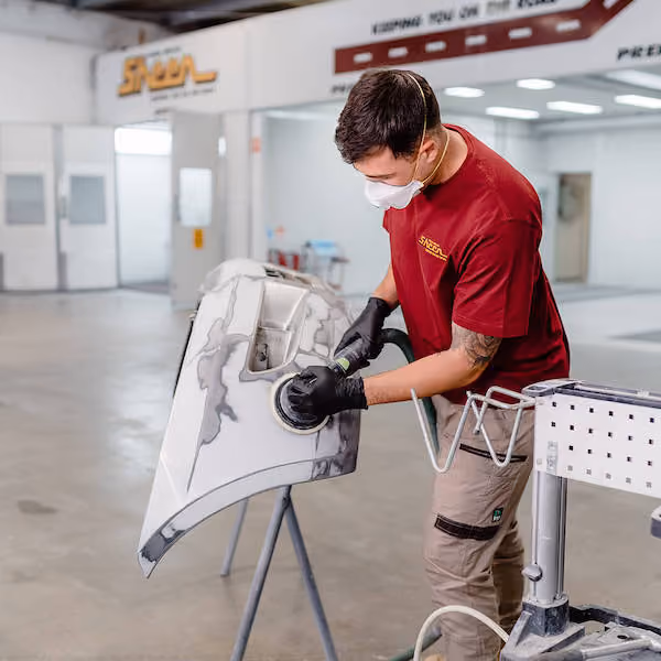 A person wearing a red shirt, black gloves, and a face mask is using a handheld power sander to smooth the surface of a car bumper in a workshop.