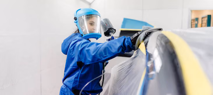 Person in blue protective suit and face shield spray painting a large object covered with plastic and yellow tape in a clean, bright workshop.